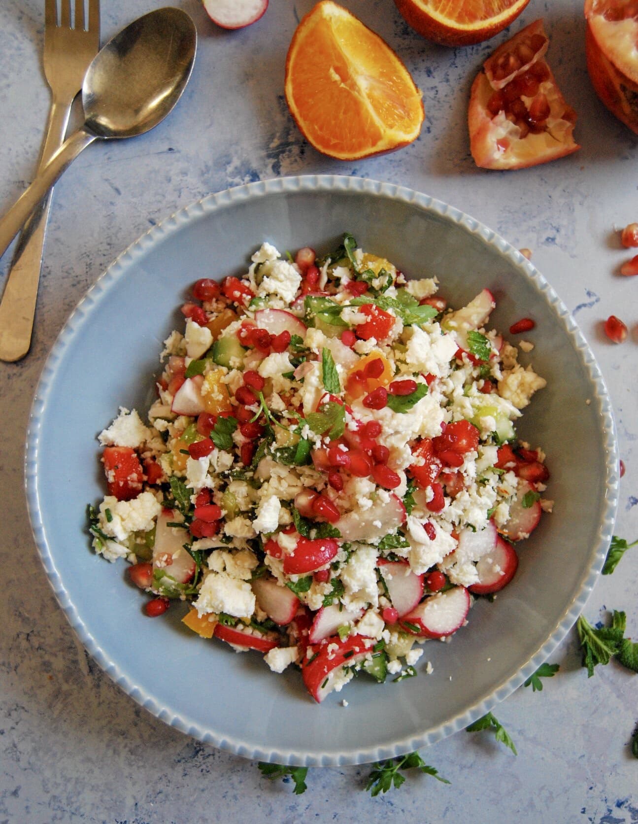 A blue bowl of Cauliflower Cous Cous Salad with radishes, pomegranate seeds and fresh green herbs.