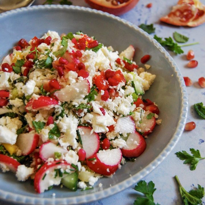 A bowl of cauliflower cous cous salad on a light blue background with pomegranate seeds and fresh herbs in the background