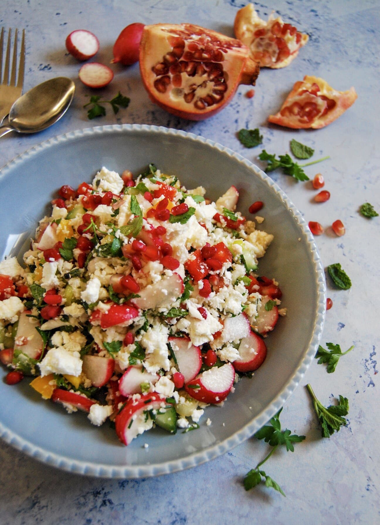 A bowl of cauliflower cous cous salad on a light blue background with pomegranate seeds and fresh herbs in the background