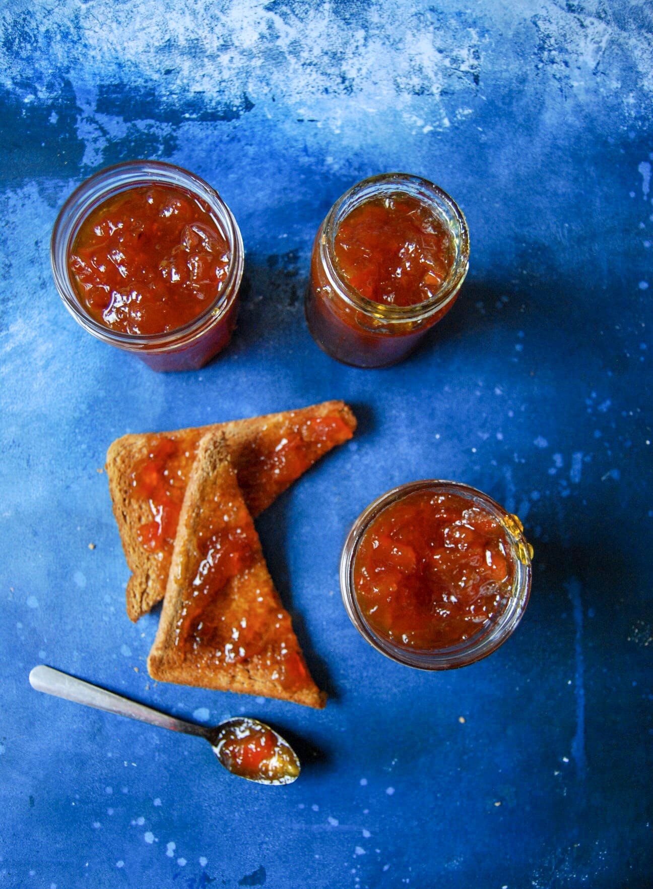 Three jars of orange marmalade, a slice of toast cut into triangles and spread with marmalade on a blue background and a silver spoon covered in marmalade.