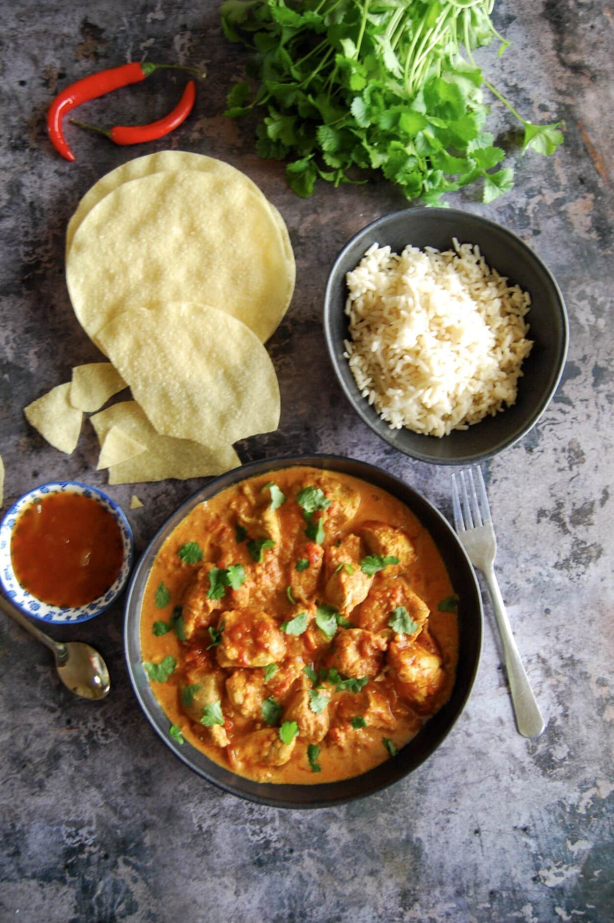 A flat lay photo of a bowl of chicken curry, poppadoms and bowls of mango chutney and rice. Red chilies and a large bunch of fresh coriander can be seen in the top corner of the photo.