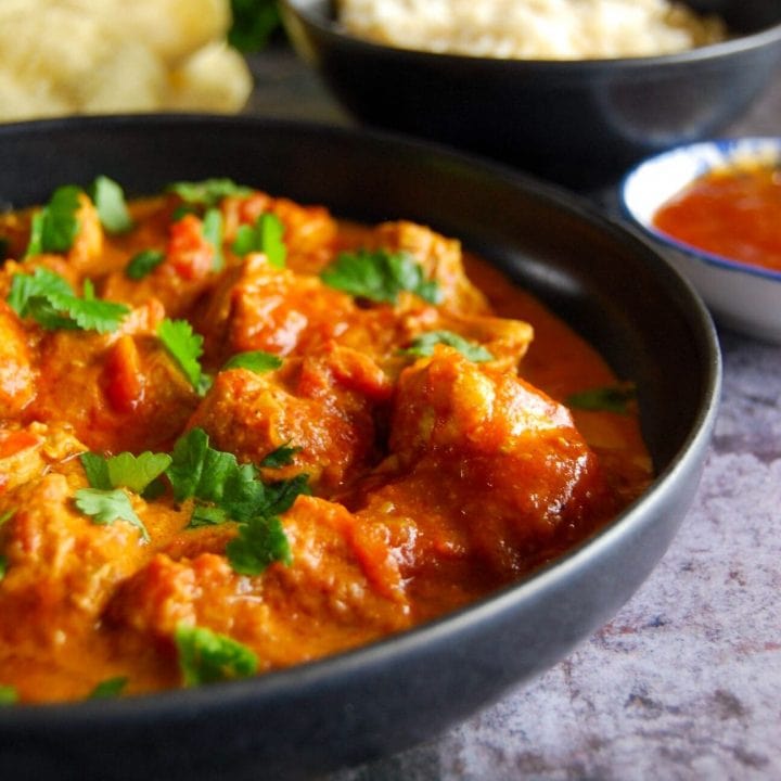 A bowl of creamy butter chicken with rice, poppadoms and mango chutney in the background