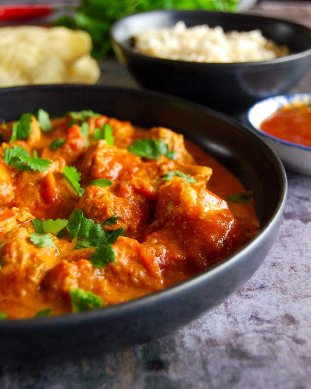 A bowl of creamy butter chicken curry with rice, poppadoms and mango chutney in the background.