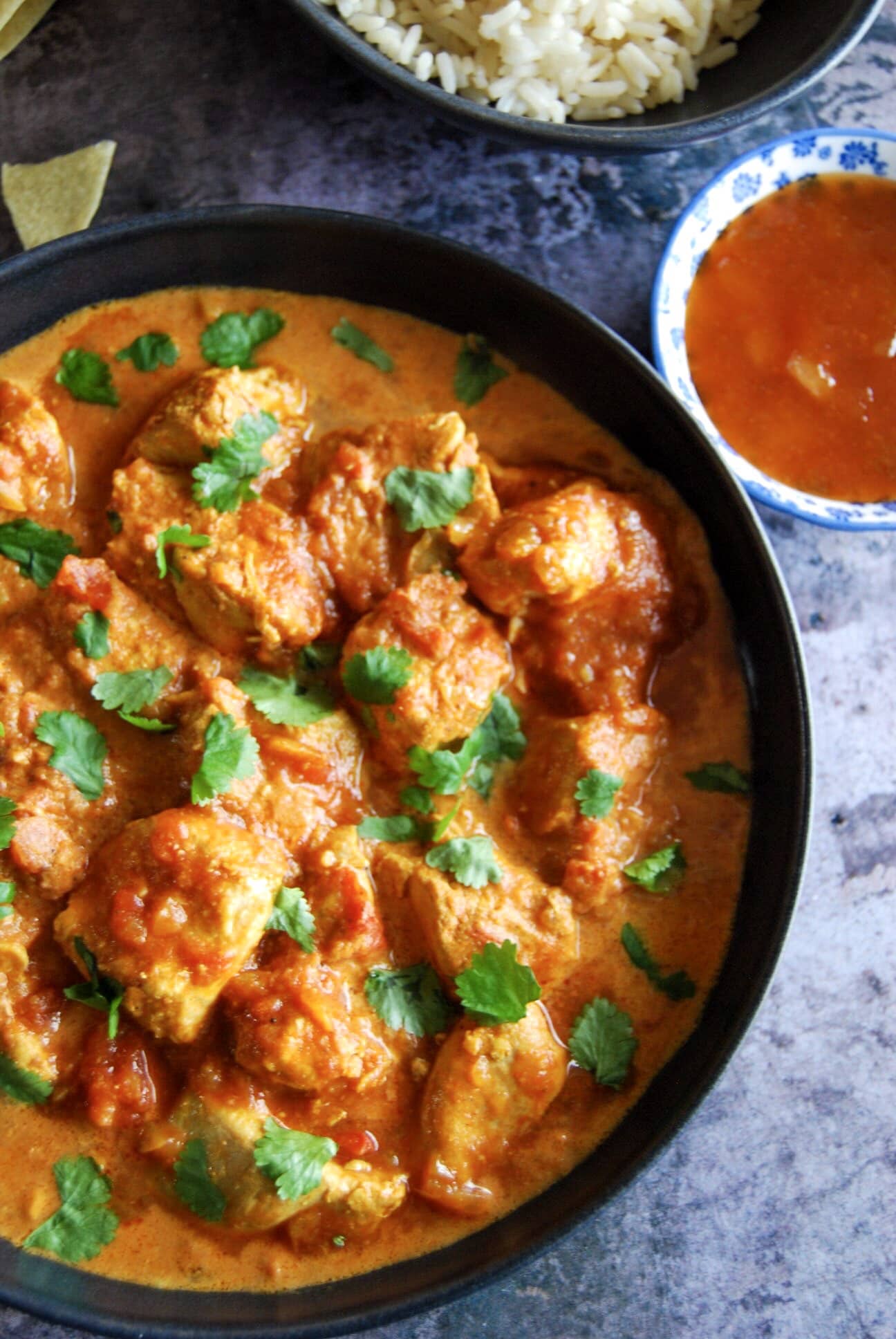 A bowl of chicken curry sprinkled with fresh coriander. Bowls of rice and mango chutney can be partially seen in the top corner.