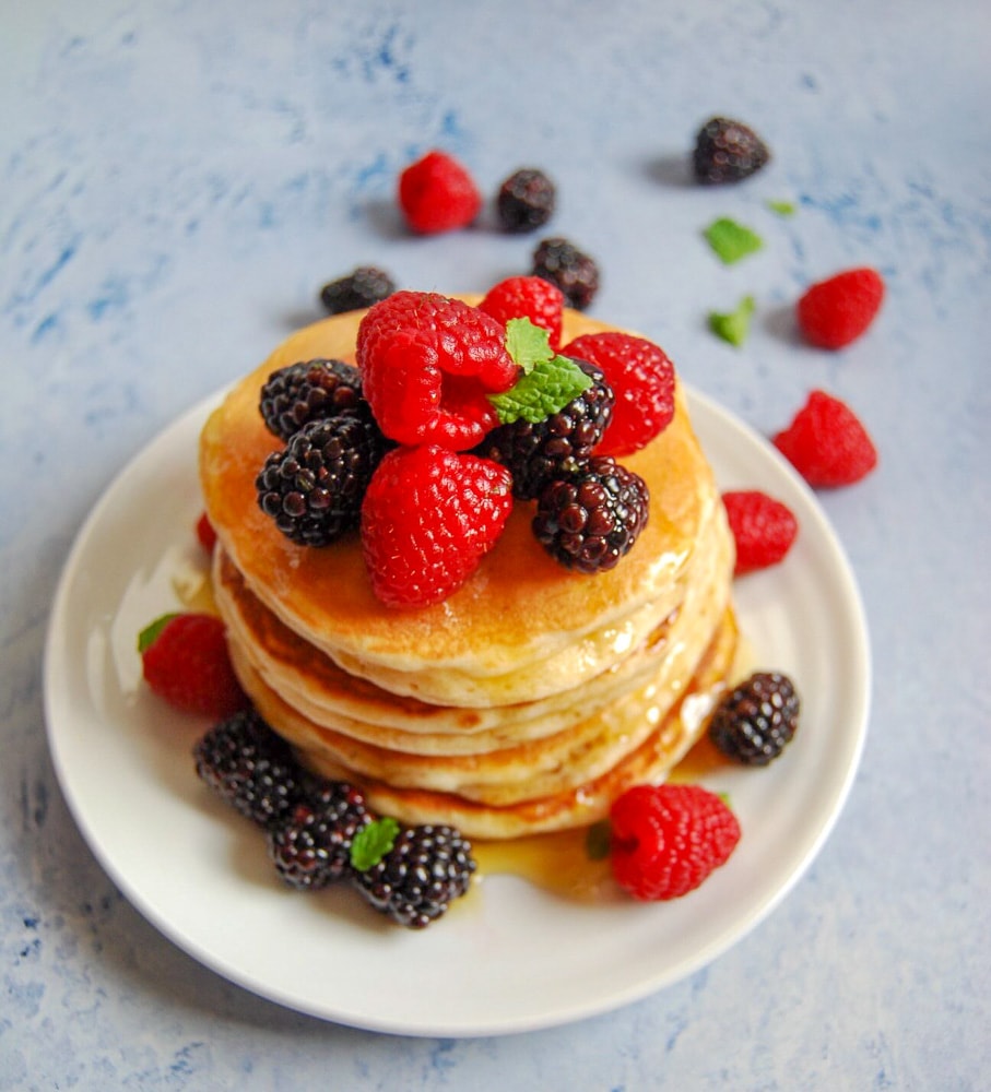 A stack of American style pancakes on a white plate topped with fresh berries and drizzled with maple syrup. 