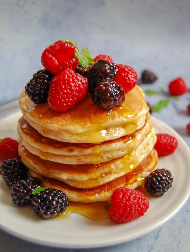 A stack of American pancakes topped with fresh berries and dripping in maple syrup.