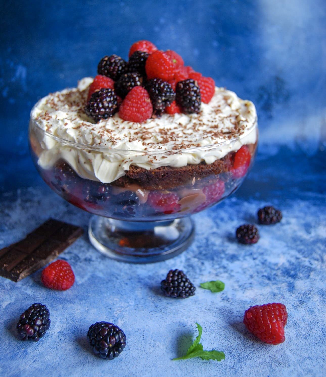 A chocolate brownie trifle topped with raspberries and blackberries in a glass bowl/sky blue backdrop