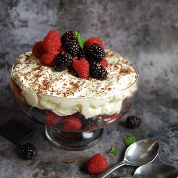 A bowl of chocolate brownie berry trifle with brownies, chocolate custard, raspberries, blackberries and whipped cream topped with chocolate shavings and more berries.