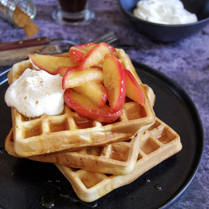 A stack of buttermilk cinnamon waffles, topped with caramelised apples and Greek yoghurt on a black plate and a dark grey background. A cup of coffee and a pot of cinnamon can be seen in the background.