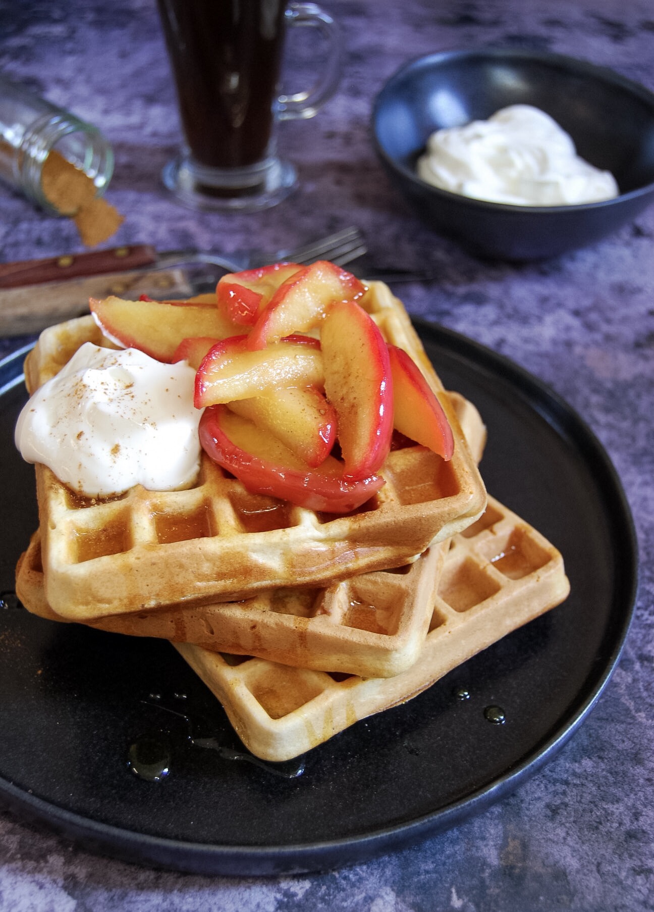 A black plate of a stack of three waffles topped with vanilla apples and greek yoghurt, a knife and fork, a latte mug of coffee and a small bowl of greek yoghurt on a grey background