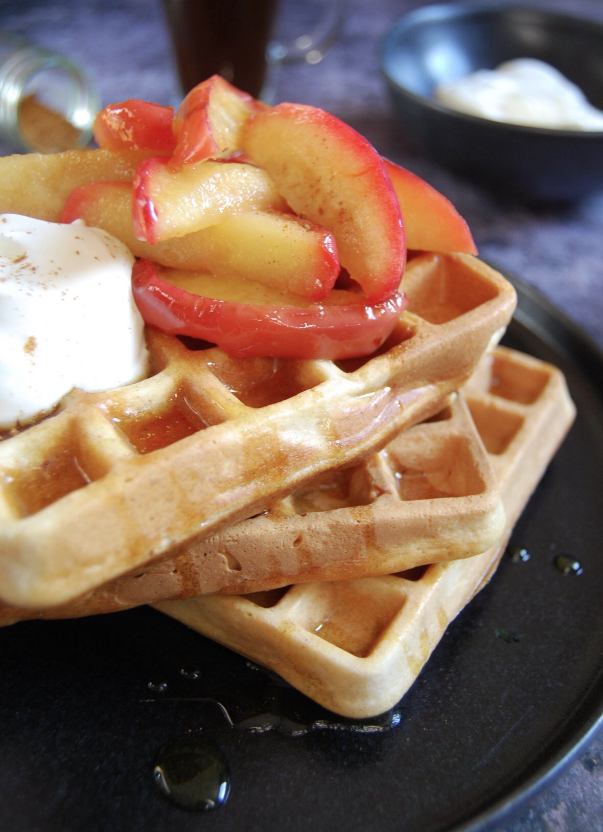 A close up photo of a stack of three waffles with maple syrup, pink apples and greek yoghurt