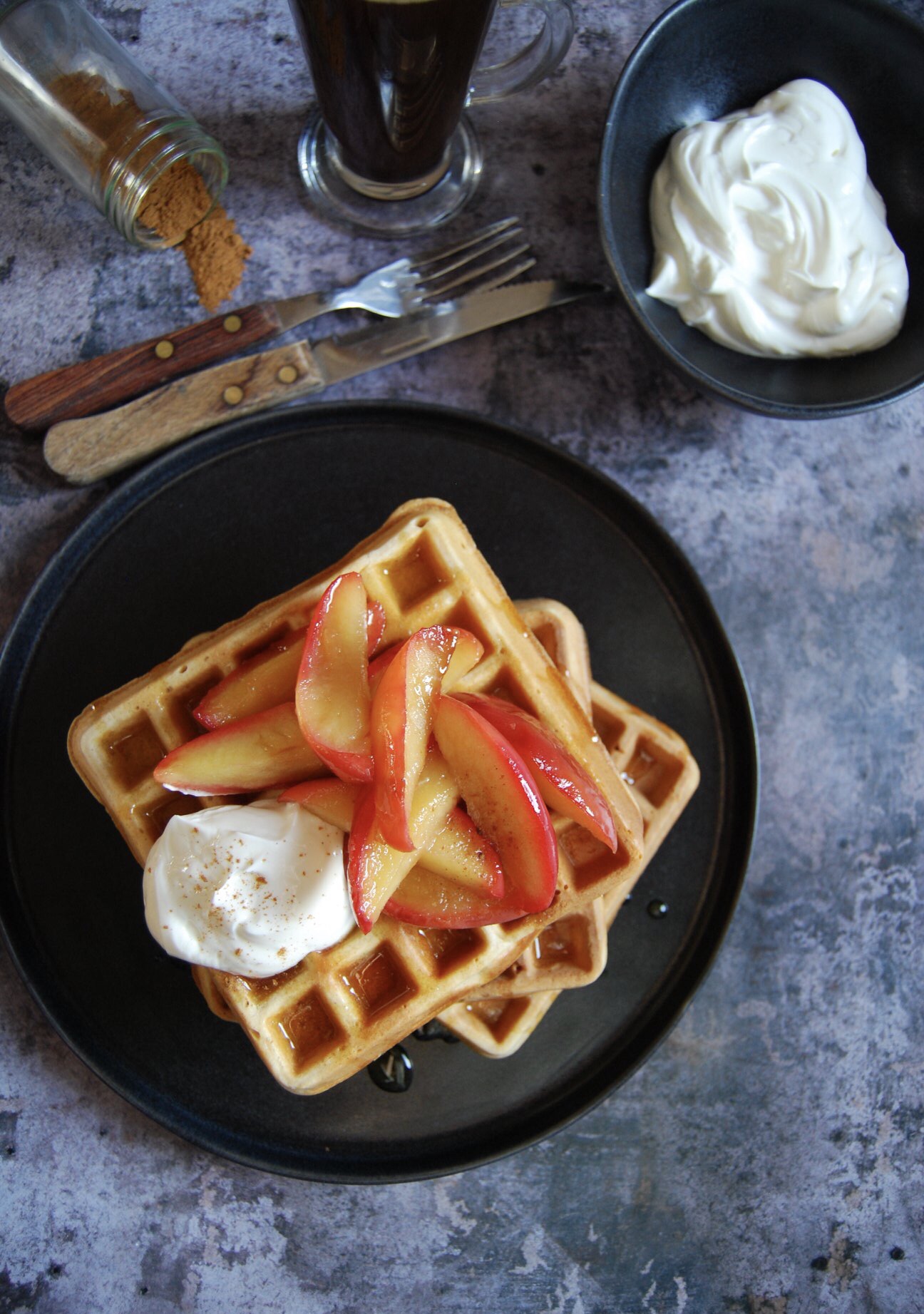 A black plate of waffles topped with apples and greek yoghurt, a wooden handled knife and fork, an upturned pot of cinnamon and a small black bowl of greek yoghurt on a grey background