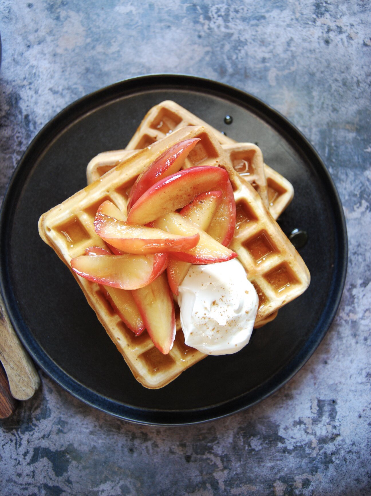 A black plate of waffles topped with pink apples and greek yoghurt on a grey background