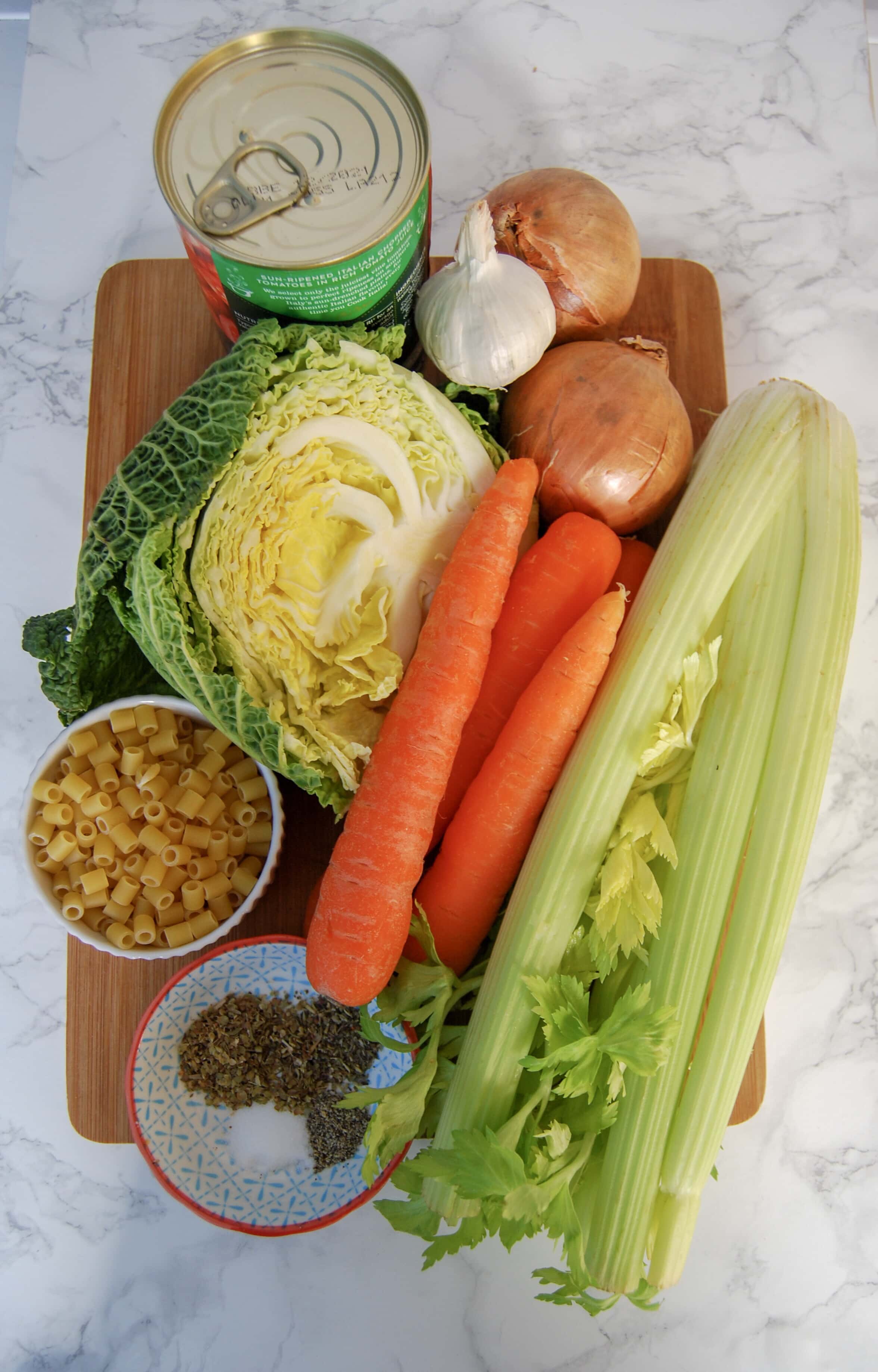 a bunch of celery, carrots, cabbage, onions, garlic, a tin of tomatoes, a bowl of pasta and a little pot of salt and pepper on a wooden board