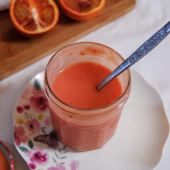 A jar of blood orange curd on a white plate with pink flowers. Blood oranges on a wooden board are in the background.