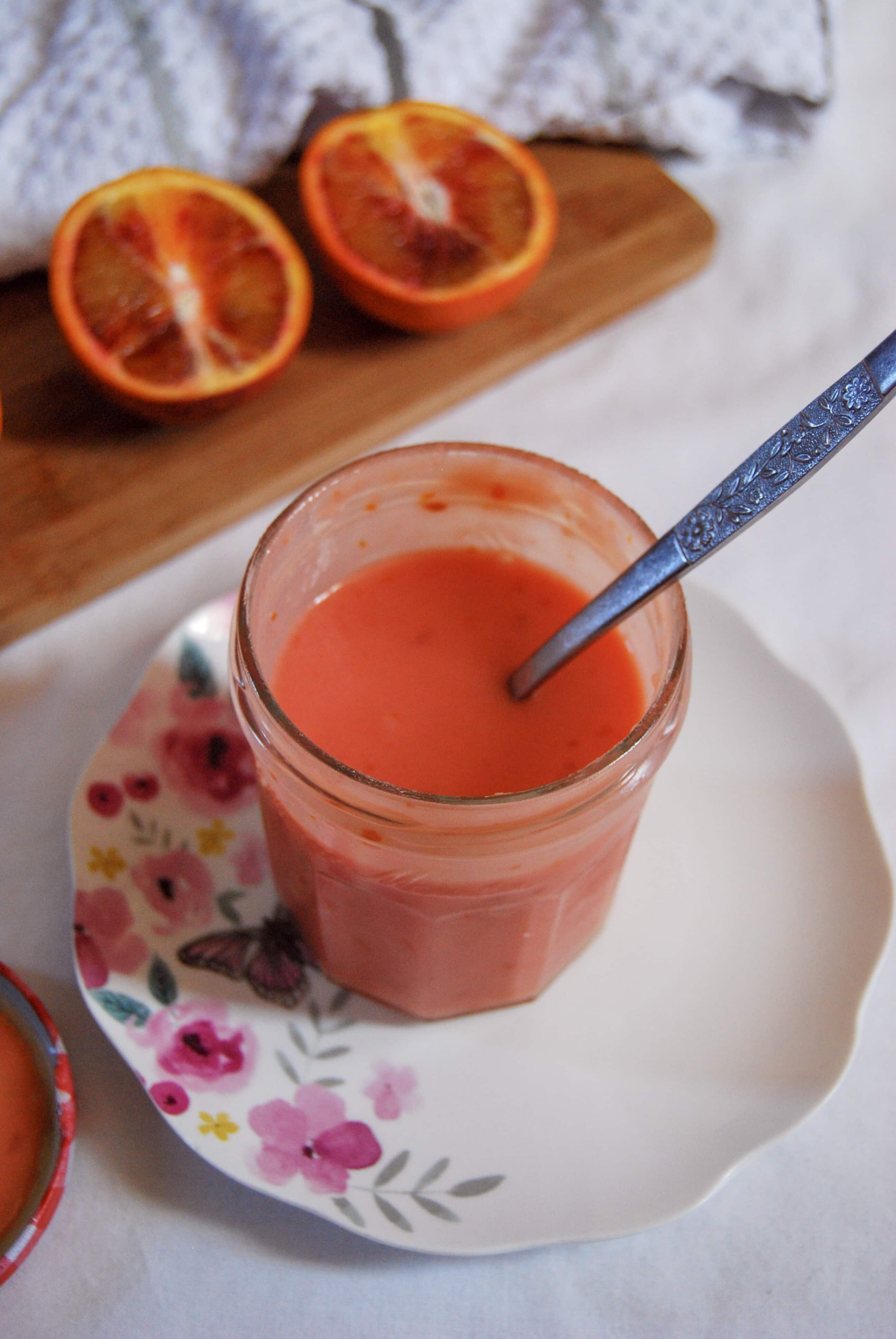 An open jar of blood orange curd on a floral plate. Halved oranges can be seen in the background. 