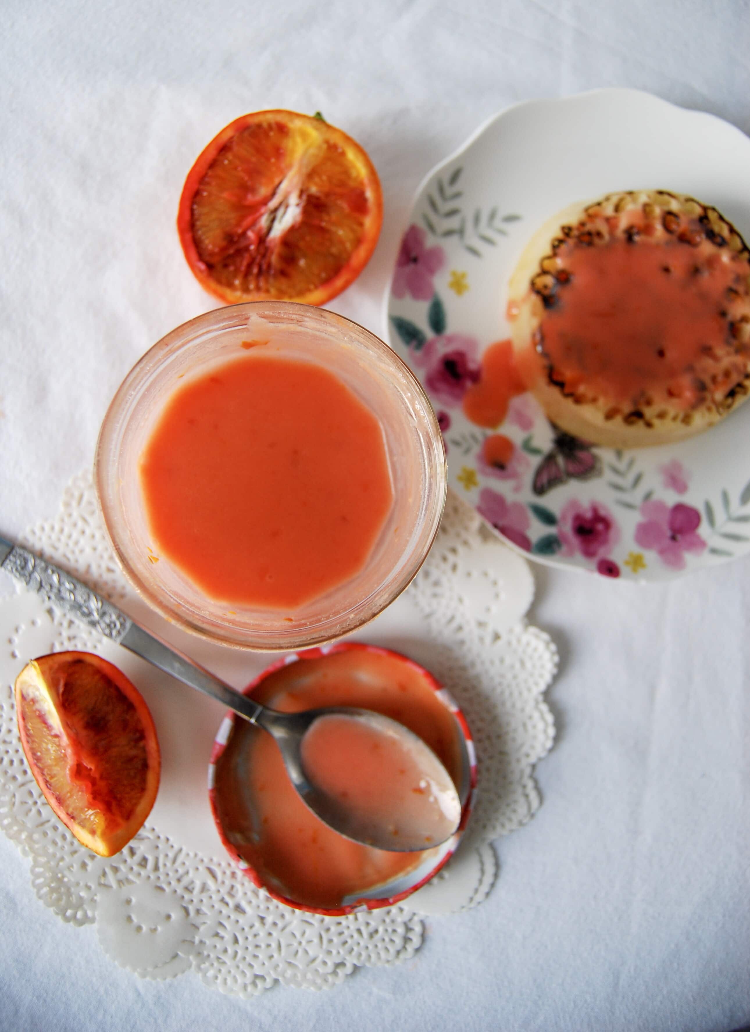 A flat lay photo of an open jar of orange curd sitting on a white paper doily, a crumpet spread with orange curd on a floral plate, a spoon resting on the jar lid and blood orange slices. 