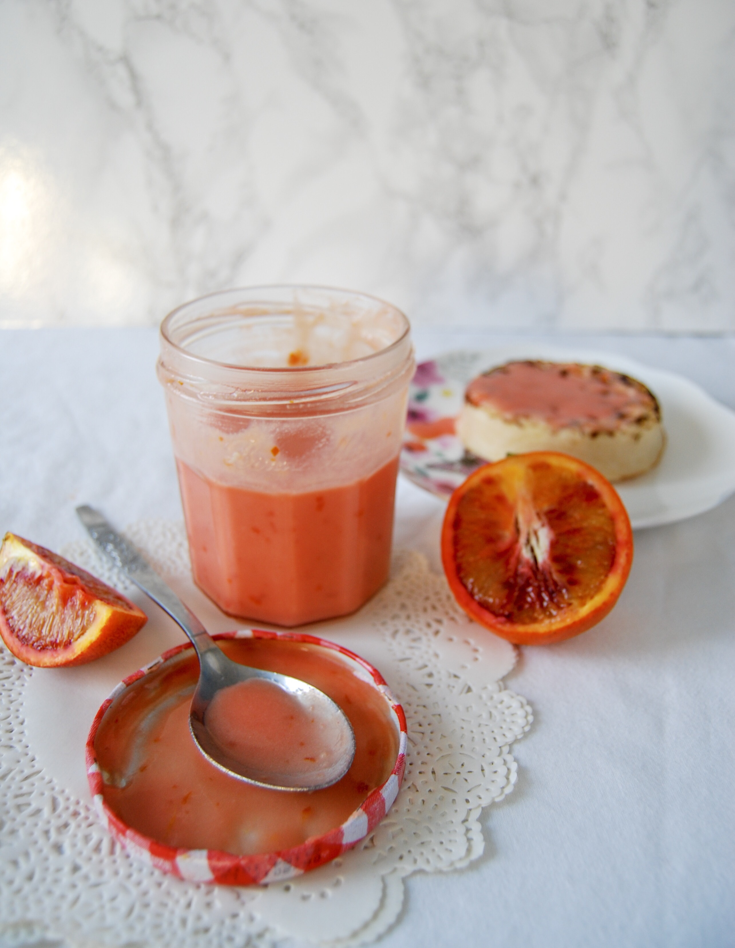 A jar of blood orange curd with a spoon resting on the jar lid. A crumpet spread with the curd can also be seen on the right hand side. 