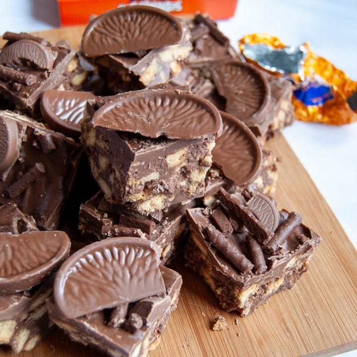 Squares of no bake chocolate orange traybake topped with matchmakers and Terry's chocolate orange on a wooden board.