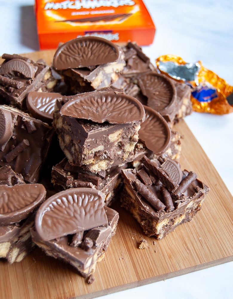 Squares of no bake chocolate orange traybake topped with matchmakers and Terry's chocolate orange on a wooden board.