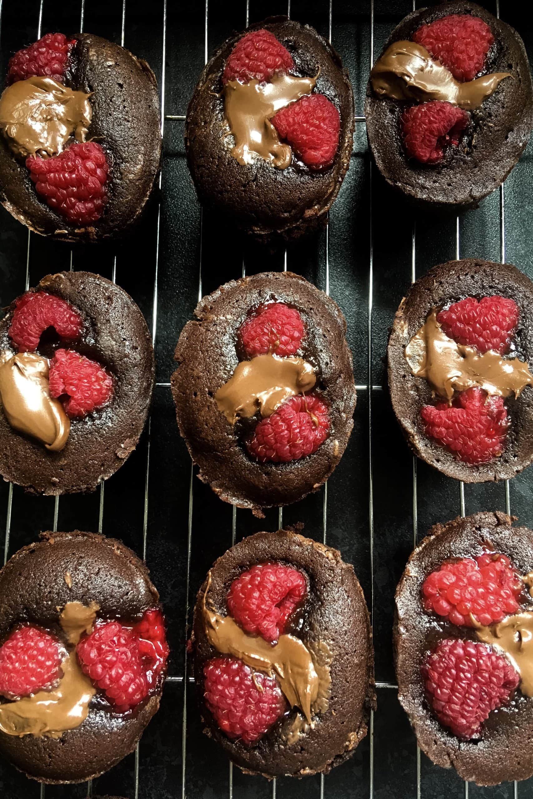 Nine little round chocolate cakes topped with fresh raspberries and hazelnut chocolate spread on a sliver wire rack
