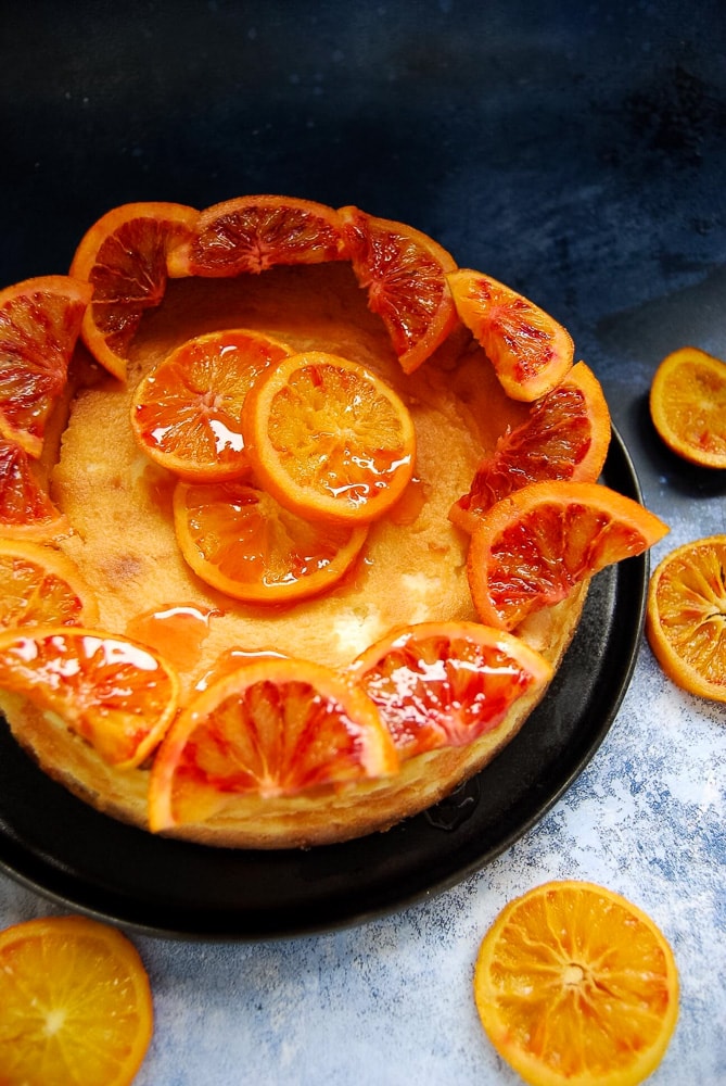A baked cheesecake decorated with candied blood orange slices on a black plate and blue background.