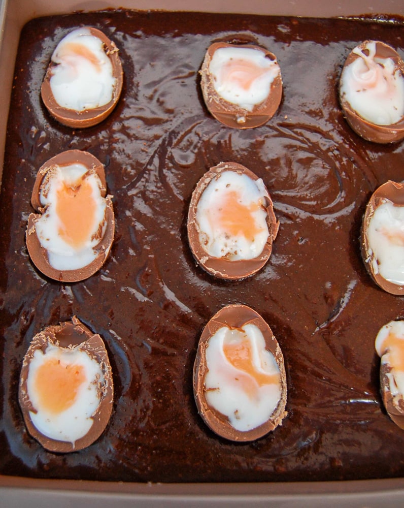 A close up photo of a slab of unbaked creme egg brownies. The creme eggs have been sliced in half and placed on top of the brownie batter.