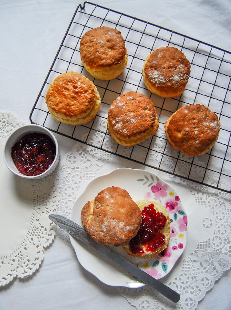 Freshly baked scones on a wire rack and a scone with butter and jam on a pretty floral plate.