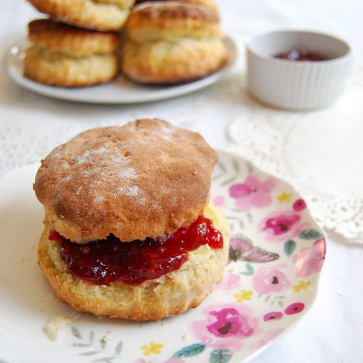 A scone filled with butter and strawberry jam on a pretty floral plate on a white tablecloth. Another plate of scones and a white pot of jam can be seen in the background.