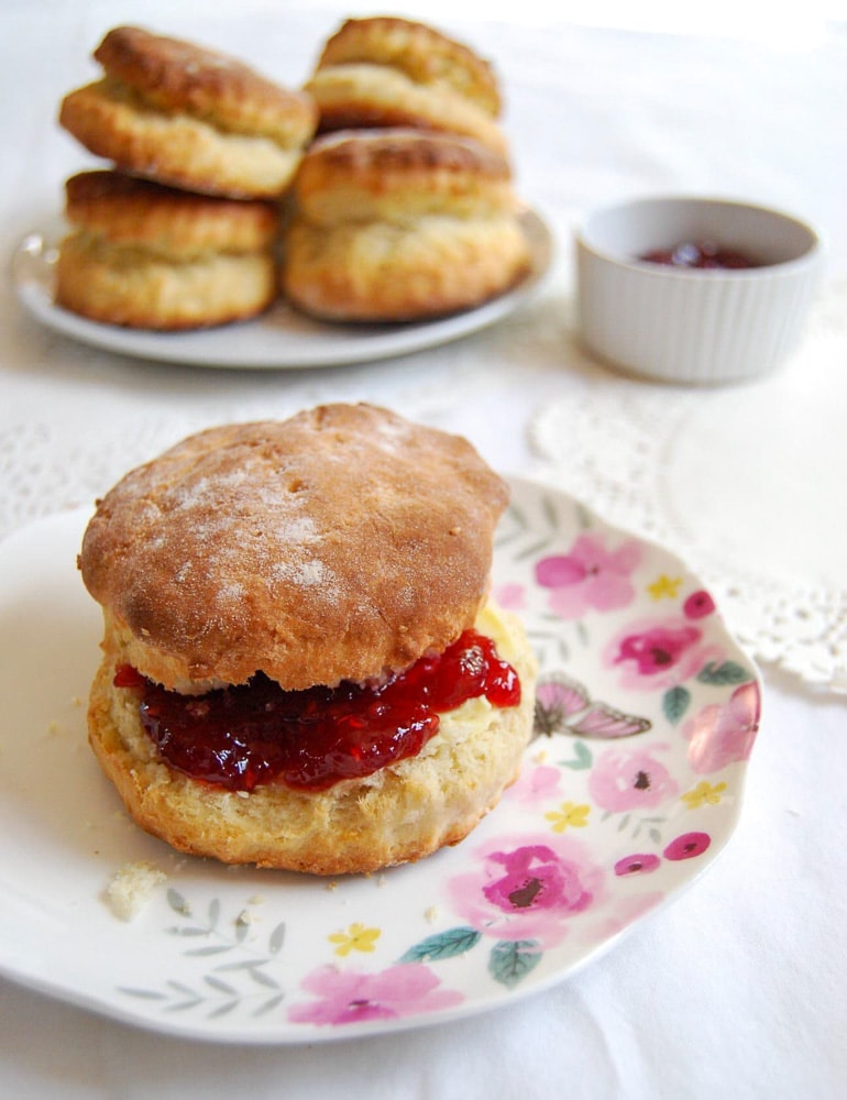 A scone filled with butter and strawberry jam on a pretty floral plate on a white tablecloth. Another plate of scones and a white pot of jam can be seen in the background.