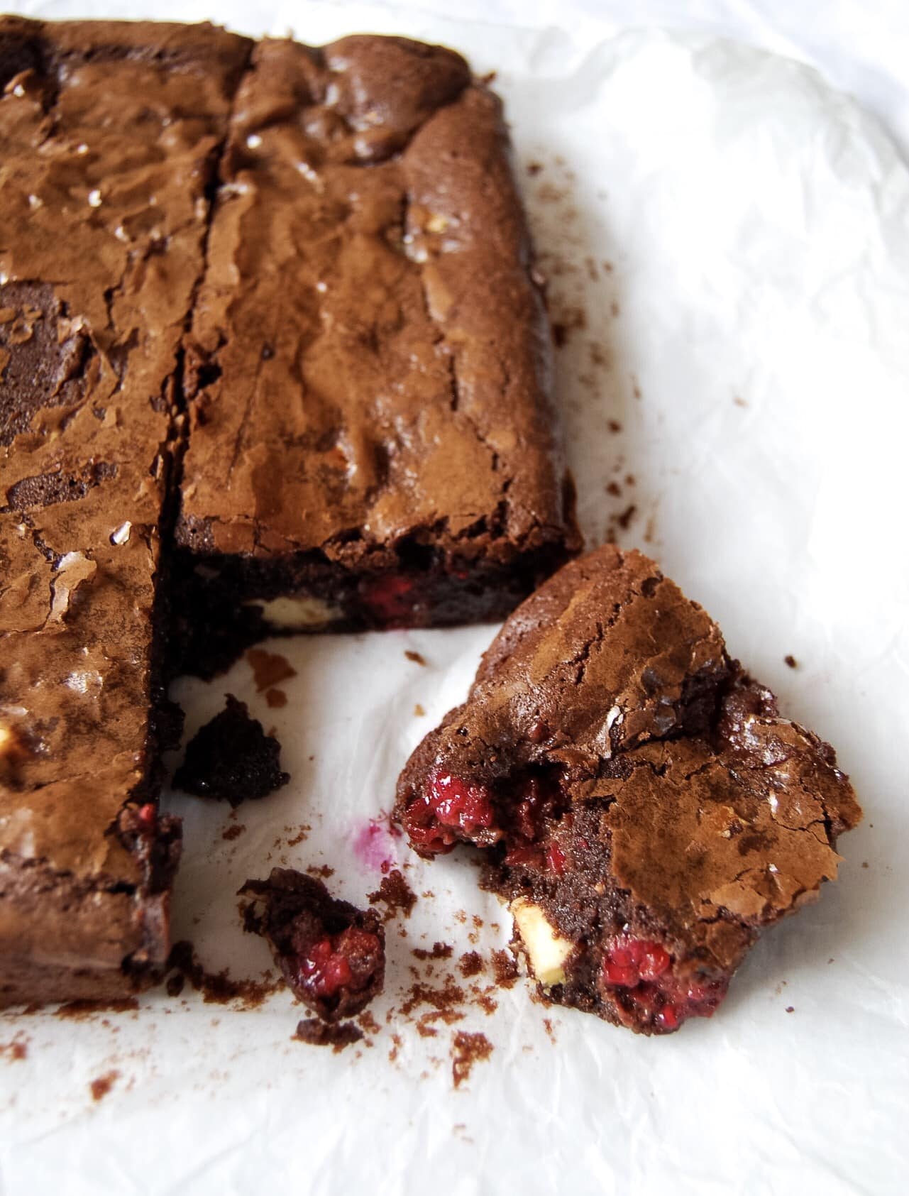A chocolate brownie with fresh raspberries and white chocolate chunks on a piece of baking parchment.