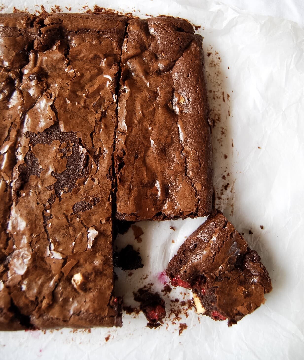 A large chocolate and raspberry brownie with a slice cut out on a sheet of baking parchment.