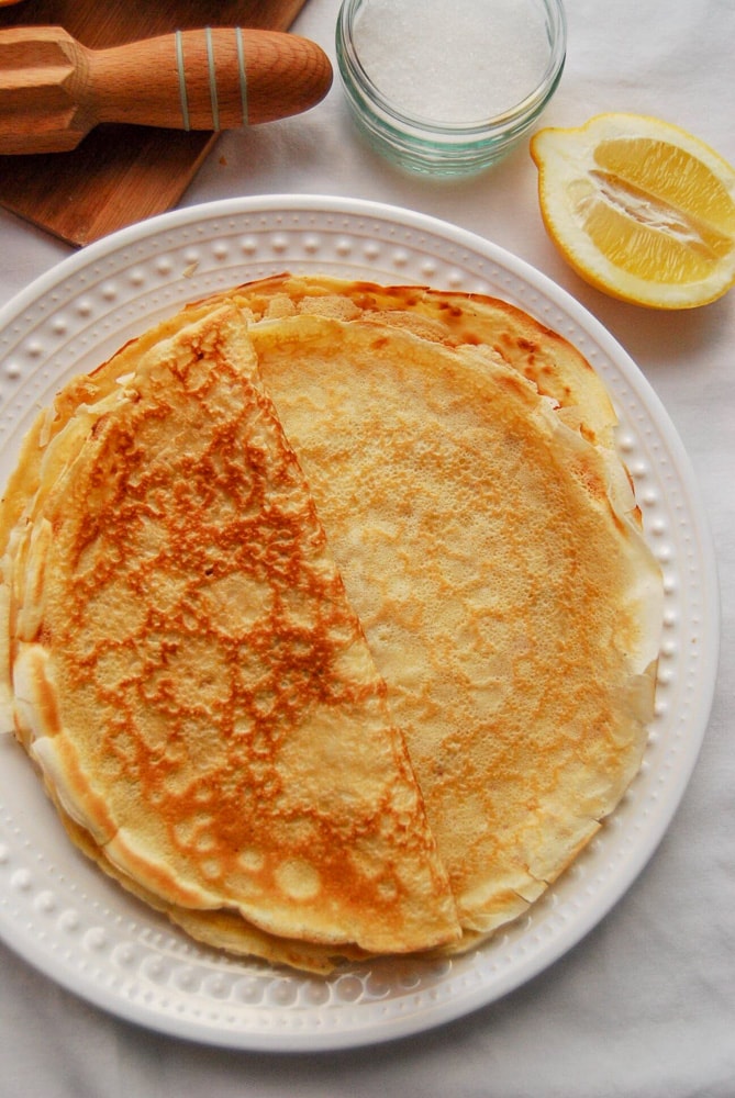 A flatlay photo of a stack of golden pancakes on a white plate. A cut lemon half and a lemon juice squeezer can be seen at the top of the picture.