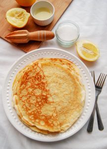 A flatlay photo of a plate of freshly made pancakes. A knife and fork, squeezed lemon halves and a wooden lemon reamer can also be seen in the picture.