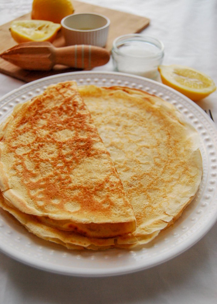 A stack of freshly made pancakes on a white plate. A lemon reamer and halved lemons can be seen in the background.