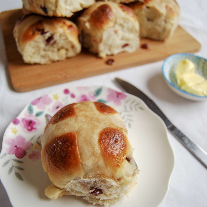 A orange, cranberry and white chocolate hot cross bun on a pretty floral plate. More hot cross buns piled up on a wooden board are in the background.