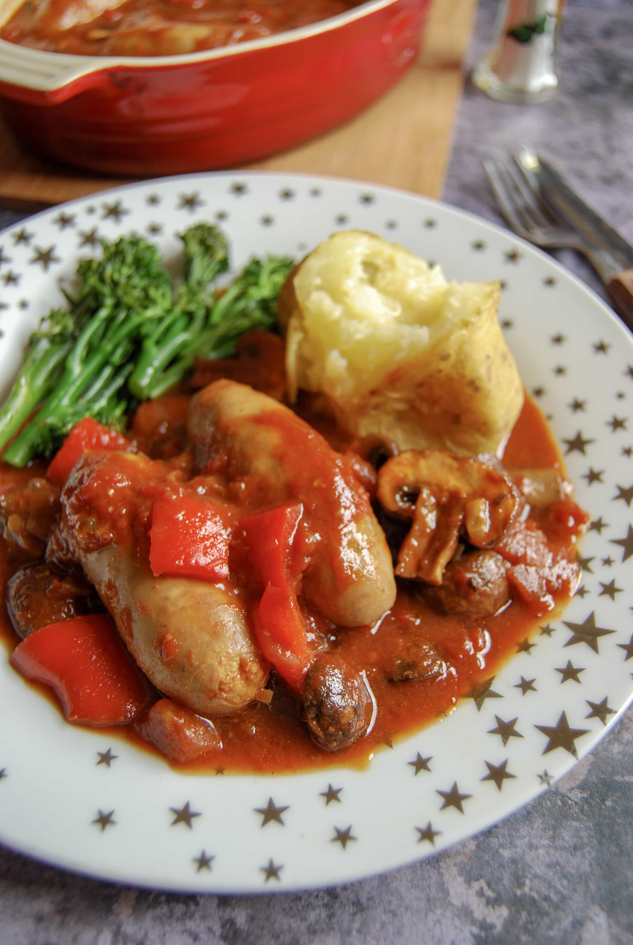 A sausage casserole with red peppers and mushrooms, baked potato and broccoli on a white and gold star plate. 