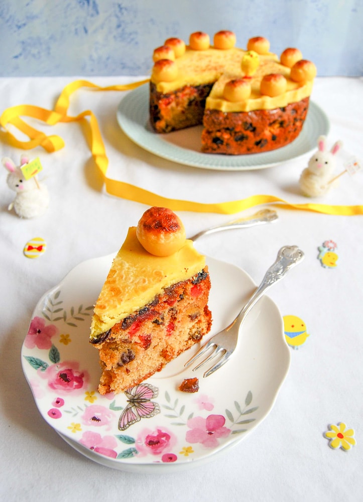 A slice of Easter Simnel fruit cake with with a layer of marzipan and a burnished golden marzipan ball on a floral plate with a silver cake fork.  A marzipan topped fruit cake on a plate in the background along with an Easter display of little rabbit figures, yellow ribbon and Easter stickers. 