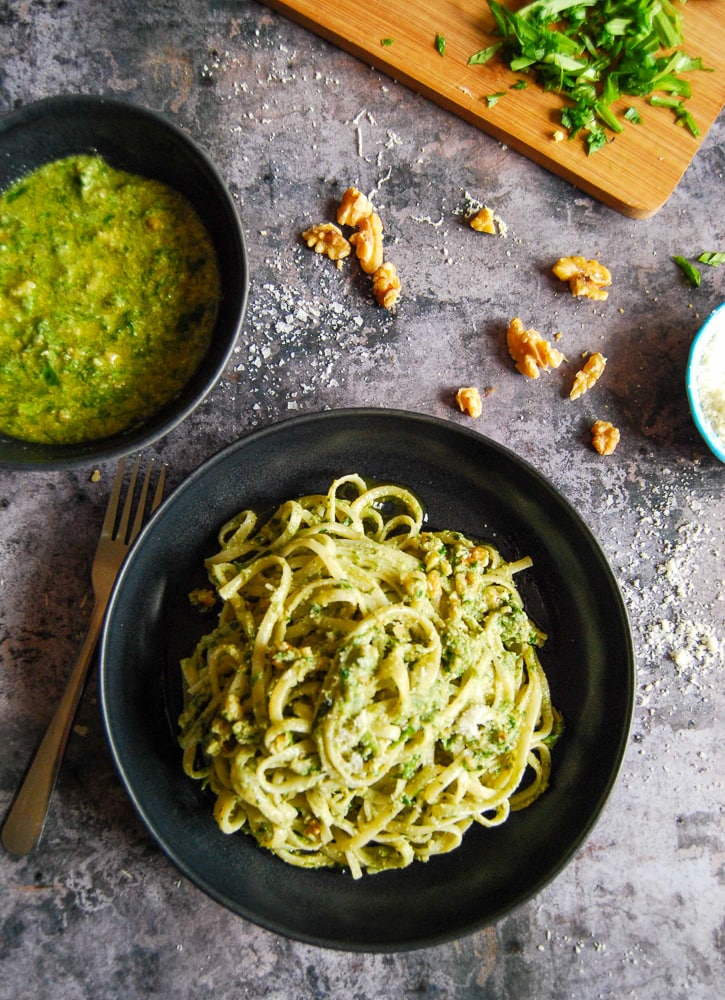 A black bowl of spaghetti with wild garlic pesto and parmesan cheese. A bowl of pasta, broken walnuts, parmesan cheese and a chopping board with basil is also in the picture.