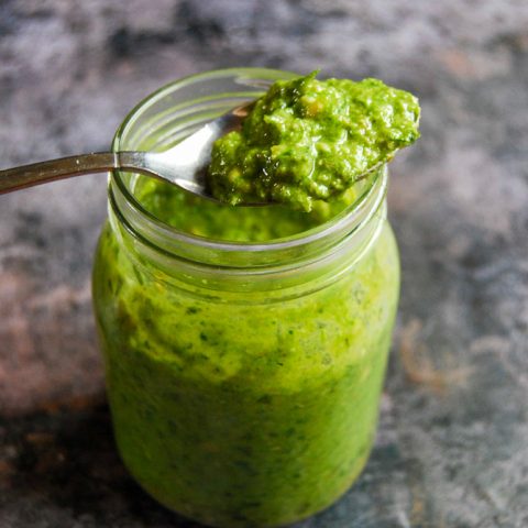 A jar of wild garlic pesto on a grey background. A spoon of pesto is resting on the jar.