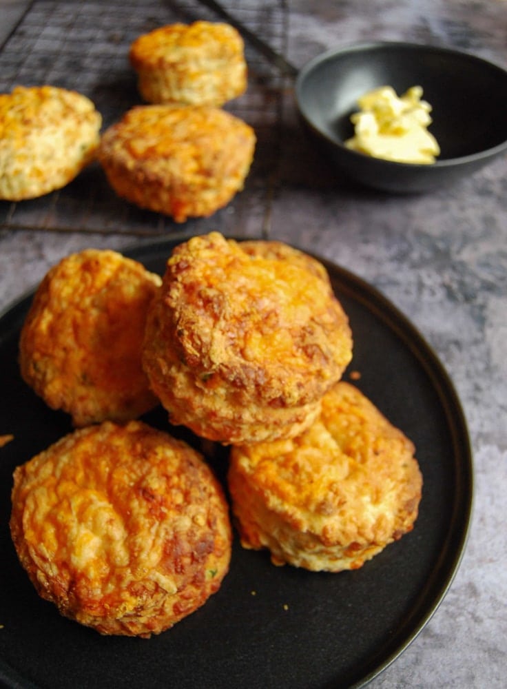 A pile of cheese scones on a black plate. A black bowl of butter and more scones on a wire cooling rack sits in the background.