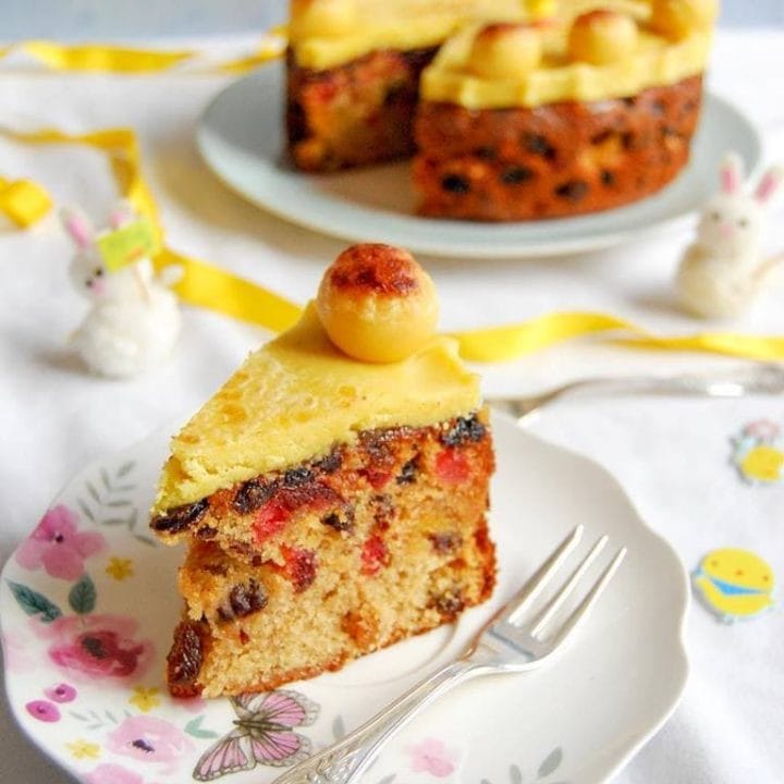 A Cut slice of Easter Simnel Cake topped with golden marzipan on a pretty floral plate. A large Simel cake sits in the background.