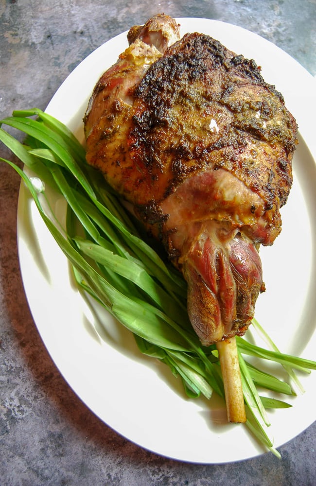 A leg of roast lamb on a white plate with freshly picked wild garlic leaves.