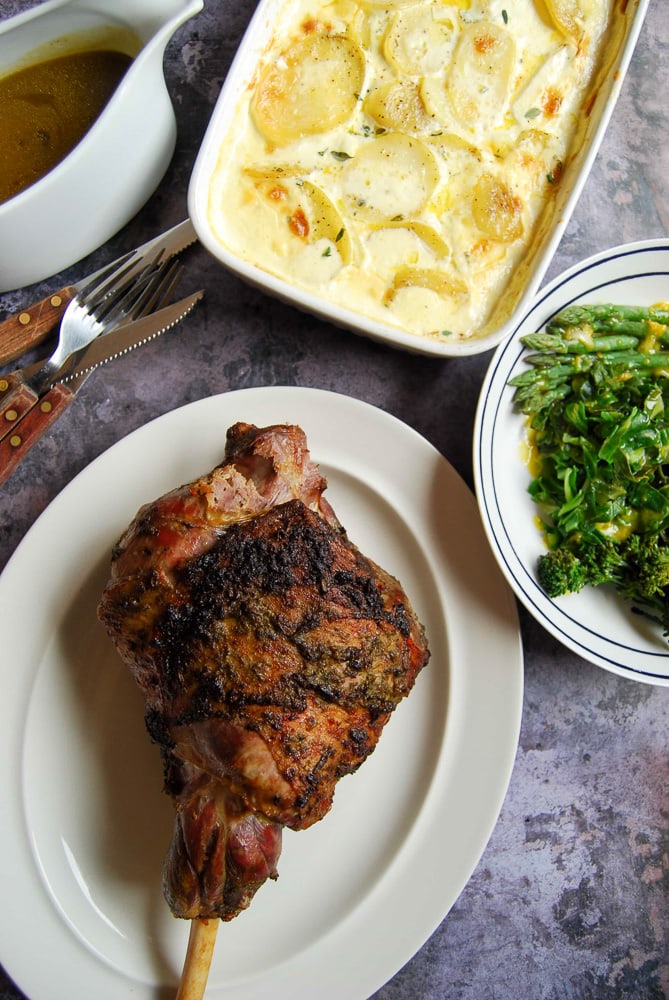 A leg of roast lamb with wild garlic and rosemary on a white plate with potato grain, Spring greens and a jug of gravy on the side.