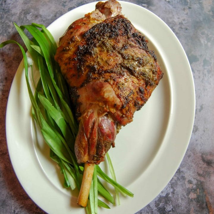 A flatlay photo of easy roast lamb with wild garlic and rosemary on a grey background. The roast lamb is sitting on a bed of wild garlic leaves.