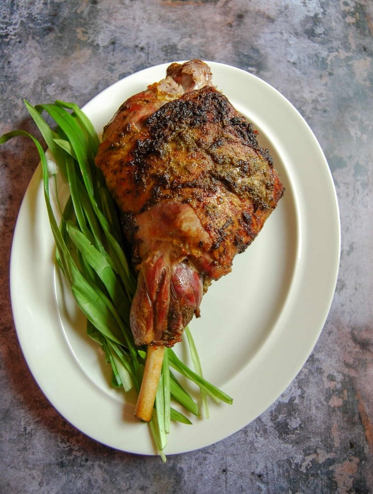 A leg of roast lamb on a white plate with freshly picked wild garlic leaves.