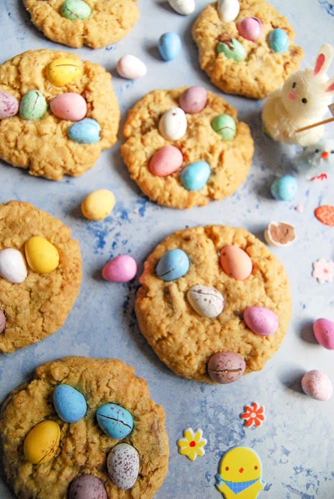 A flatlay picture of Mini Egg Cookies on a light pastel blue background. Mini pastel coloured chocolate eggs sit in between the cookies along with a cute little Easter bunny.