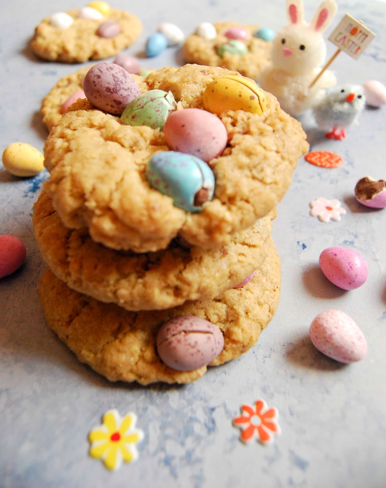 A stack of Mini Egg Cookies on a light pastel blue background with mini pastel chocolate eggs in the background
