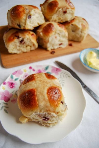 A hot cross bun on a white and pink plate. A pile of hot cross buns can be seen in the background on a wooden board.
