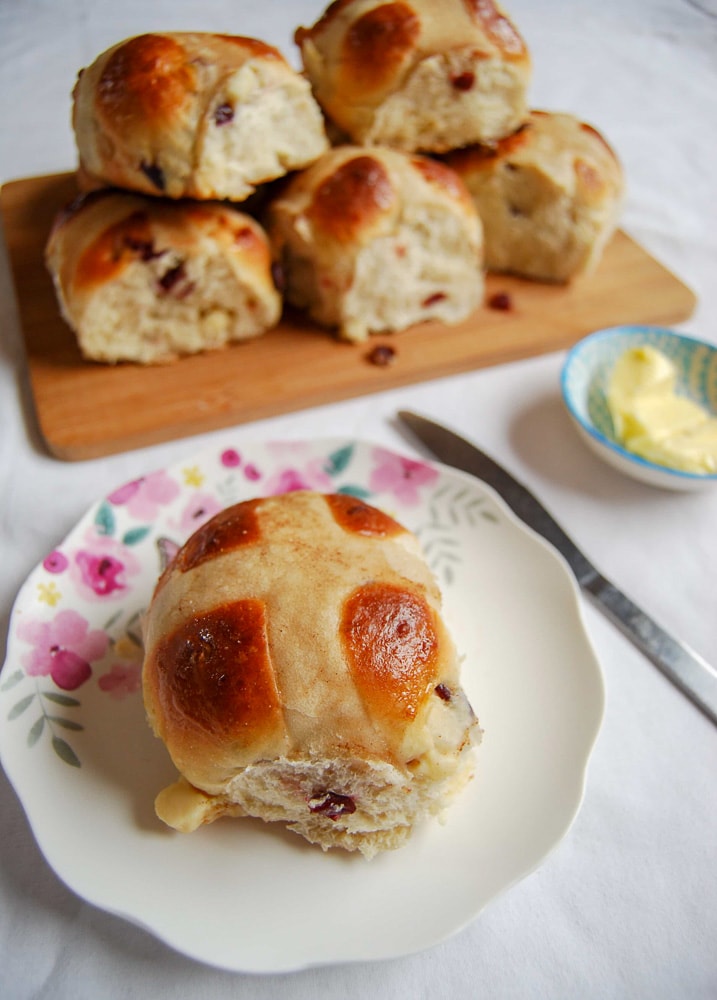 A hot cross bun on a floral plate with a small pot of butter and a butter knife. A board with more hot cross buns can be seen in the background.